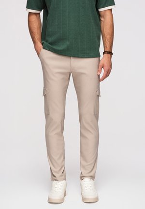 Mężczyzna w beżowych spodniach typu cargo, białych trampkach i ciemnozielonym krótko rękawowym swetrze, z rękami swobodnie opuszczonymi wzdłuż ciała.