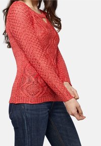 Czerwony, teksturowany, dziany sweter z długim rękawem, z dekoltem w kształcie łezki i wzorem warkocza, wykończony ściągaczami na mankietach i dole. Noszony z ciemnoniebieskimi dżinsami.