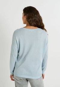 Femme aux longs cheveux bruns portant un pull léger bleu clair à manches longues et un pantalon gris clair, de dos devant un fond uni.