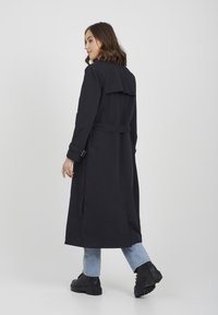 Czarny płaszcz trench z paskiem, długimi rękawami i detalem na plecach. Nosi się go na jasnoniebieskie dżinsy i czarne buty na grubej podeszwie.