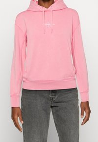 Rosa hoodie med känguruficka, ribbade ärmslut och nederkant, med en liten vit logotyp. Tillverkad av mjukt tyg med en avslappnad passform.