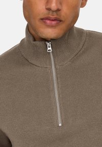 Maglione a coste marrone con colletto alto e mezza zip sul davanti. Il tessuto ha un aspetto texture, suggerendo calore e comfort.