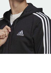 Persona che indossa una felpa nera con zip Adidas, con strisce bianche sulle maniche, parzialmente zipata, che mostra una maglietta bianca sottostante.
