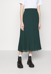 Flerad midikjol i lätt tyg i djupgrönt, med ett elastiskt midjeband och en flödande drapering, tillsammans med svarta kraftiga stövlar.