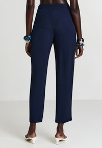 Femme portant un pantalon droit bleu marine, des mules blanches à talons, et des bracelets épais, se tenant devant un fond clair et uni.