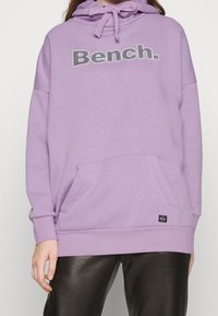 Fioletowa bluza z kapturem z kieszenią z przodu i napisem "Bench", noszona przez osobę w czarnych skórzanych spodniach, pokazana od szyi do bioder.