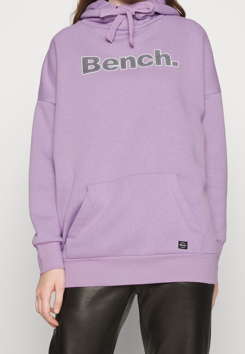 Fioletowa bluza z kapturem z kieszenią z przodu i napisem "Bench", noszona przez osobę w czarnych skórzanych spodniach, pokazana od szyi do bioder.