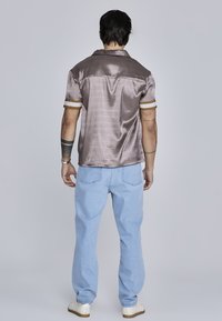 Camisa de manga corta en un brillante tejido topo con un patrón sutil, que cuenta con puños a rayas en blanco y dorado, combinada con unos jeans azul claro.