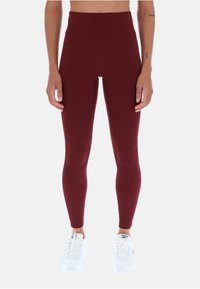 Leggings burdeos de cintura alta con una textura suave y elástica. Combinados con zapatillas deportivas blancas. Sin patrones ni acentos visibles.