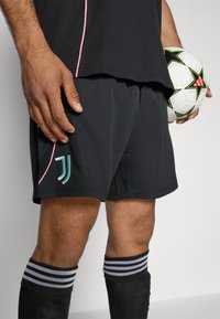 Schwarze Sportshorts mit hellrosa Paspelierung, ausgestattet mit einem türkisfarbenen Logo auf der linken Seite und schwarzen knielangen Socken mit weißen Streifen.
