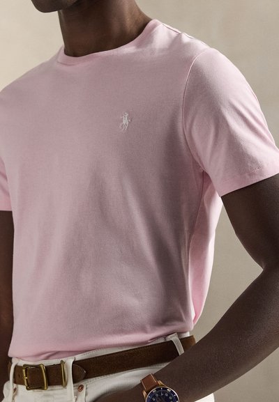 Ανοιχτό ροζ βαμβακερό t-shirt με στρογγυλή λαιμόκοψη και λευκό κεντημένο λογότυπο στο στήθος. Συνδυασμένο με καφέ ζώνη και ρολόι.