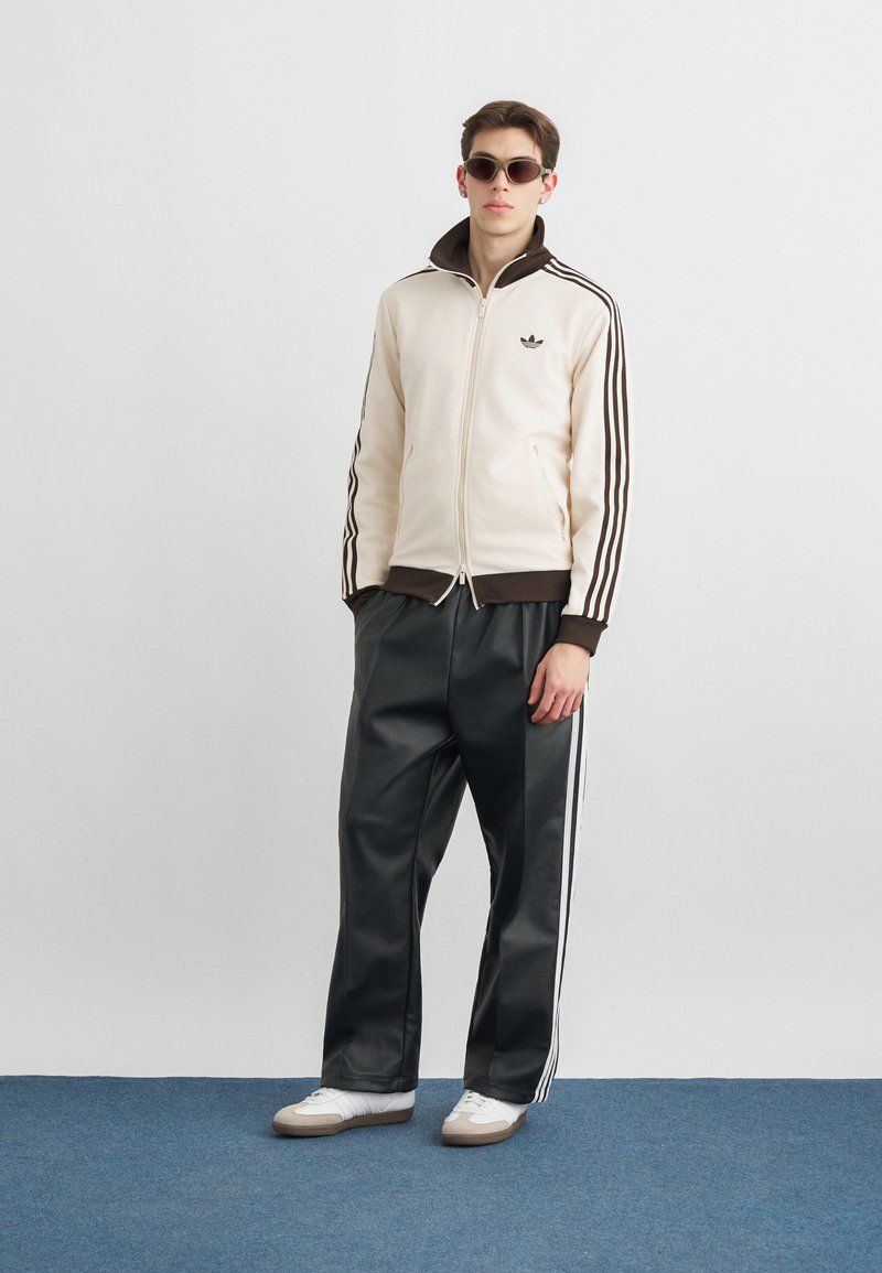 Joven con chaqueta con cremallera Adidas en crema y marrón, pantalones deportivos negros con rayas blancas, zapatillas blancas y gafas de sol marrones, de pie sobre una alfombra azul.