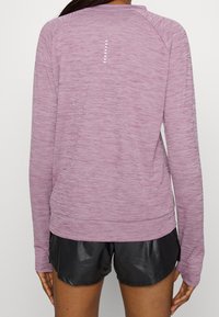 Langärmeliges Sportshirt in hellem Lila aus strukturiertem Stoff, mit reflektierenden Akzenten auf dem Rücken, kombiniert mit schwarzen Shorts.