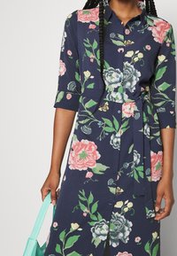 Vestito camicia floreale in tessuto blu navy con motivi di fiori rosa, verdi e blu, chiusura frontale a bottoni e dettaglio con cintura in vita. Inclusa borsa aqua.