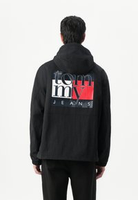 Zwarte hoodie met een gestructureerde afwerking, voorzien van een gedurfde logoprint in wit en rood over de achterkant. Elastische manchetten maken de look compleet.