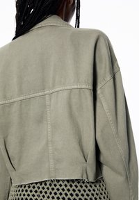 Kort jasje in olijfgroene, duurzame stof met een naad in het achterpand en een ongezoomde onderkant. Draagt over een gestructureerde zwarte nettop.