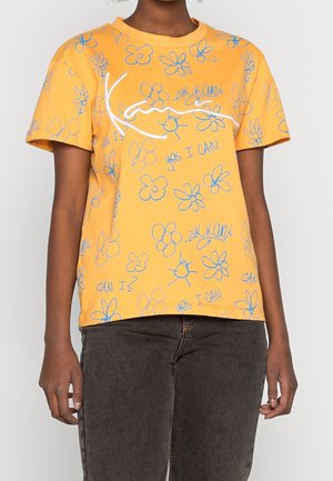 Žluté tričko s krátkým rukávem s modrými květinovými náčrty a nápisy jako "ano, dokážu to", nošené s tmavě šedými kalhotami.