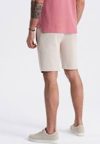 Bermudas beige para hombre, hechas de una tela suave, presentan una textura lisa y un corte relajado, combinadas con zapatillas grises claras y una camiseta rosa.