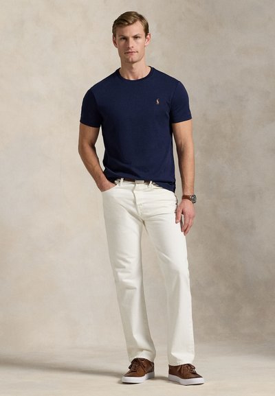 Moški v mornarsko modri majici, belih hlačah, rjavem pasu, rjavi ročni uri in rjavih supergah, stoji z eno roko v žepu.