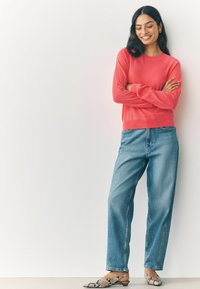 Korallfarbener Pullover mit langen Ärmeln, kombiniert mit lockeren blauen Jeans und gemusterten, flachen Schuhen. Einfaches Design vor hellem Hintergrund.