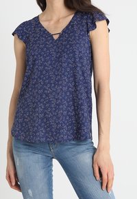 Donna che indossa una blusa corta a maniche corte blu con motivo floreale e scollo a V, abbinata a jeans chiari strappati, in piedi davanti a uno sfondo grigio chiaro uniforme.