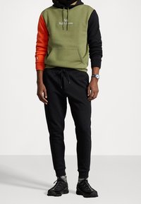 Bluza z kapturem o oliwkowozielonym korpusie, pomarańczowym rękawie i czarnym kapturze. Z ozdobnym, wyszywanym logo. Połączona z czarnymi spodniami dresowymi i czarnymi butami sportowymi.