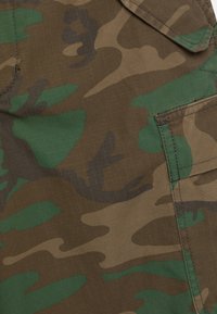 Tkanina maskująca w brązowe, zielone i beżowe wzory. Charakteryzuje się teksturowanym splotem oraz boczną kieszenią z detalami szwów.