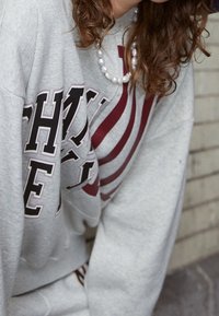 Felpa grigia con audace testo grafico in maroon scuro e design a strisce. Presenta una vestibilità comoda e una texture morbida. Collana a perle visibile.