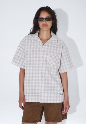 Kvinde iført sorte solbriller, en løs beige ternet kortærmet skjorte med en frontlomme og brune knælange shorts mod en ensfarvet baggrund.