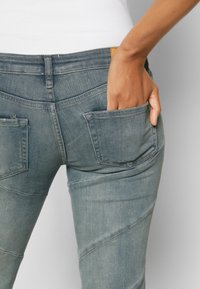 Calças de ganga azul desbotadas com um corte slim. Apresenta dois bolsos traseiros funcionais com aba, costuras visíveis e uma superfície texturizada.