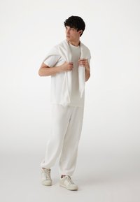 Camiseta de algodón blanca con el logo "QUE", combinada con pantalones de chándal blancos. Un suéter blanco está caído sobre los hombros. Zapatillas beiges completan el conjunto.