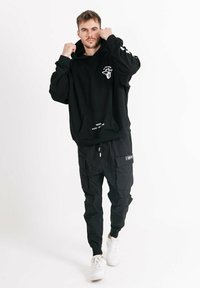 Schwarzer Kapuzenpullover mit Bauchtasche und grafischem Druck, kombiniert mit schwarzen Cargo-Hosen mit mehreren Taschen und Details. Weiße Sportschuhe.