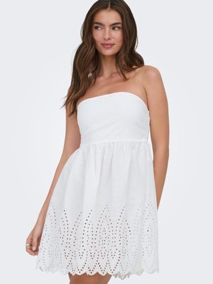 Vrouw met bruin haar in een strapless witte jurk met oogjesdetails langs de geschulpte zoom, kijkt naar links.