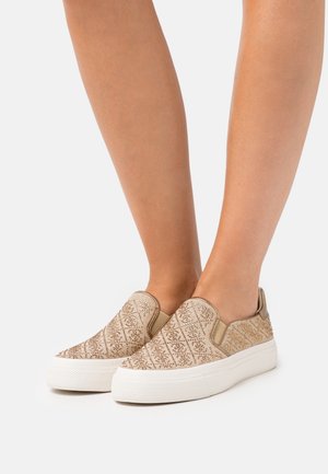 Jambes portant des baskets beige à enfiler avec un motif en losanges texturé et des semelles épaisses en caoutchouc blanc sur un fond blanc.