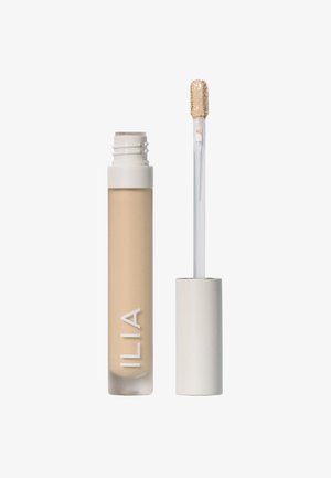 Correcteur ILIA dans un tube transparent avec un produit beige visible. Il présente un capuchon blanc et un applicateur transparent avec un embout texturé.