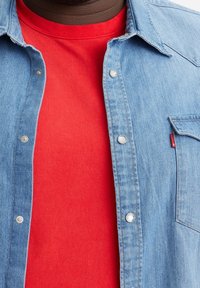 Zbliżenie na tors w jasnoczerwonej koszuli pod rozpiętą jasnoniebieską kurtką dżinsową z metalowymi napami.