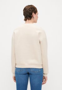 Ανοιχτό μπεζ φούτερ με στρογγυλή λαιμόκοψη, χαμηλούς ώμους και άνετη εφαρμογή. Μαλακό, λείο ύφασμα, σε συνδυασμό με μπλε skinny τζιν.