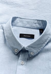 Camicia azzurra pallido a maniche corte con colletto a punta, realizzata in un tessuto texturizzato. Presenta due bottoni bianchi e un'etichetta nera.