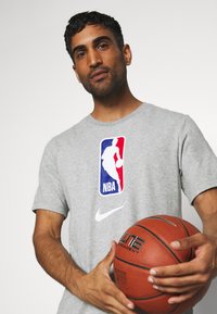 Grå kortærmet t-shirt med NBA-logo og Nike-swoosh, der holder en basketball, med en glat bomuldsstruktur og afslappet pasform.