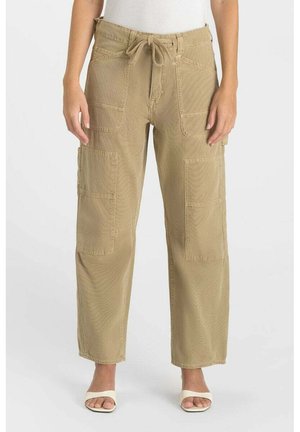 Vrouw draagt beige cargobroek met wijde pijpen, trekkoortaille en meerdere grote voorzakken, gecombineerd met witte sandalen met open neus.