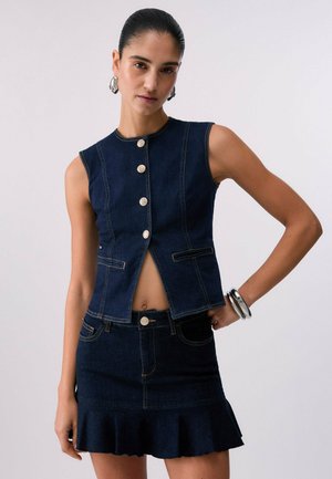 Donna con capelli pettinati all'indietro indossa un top in denim scuro senza maniche con bottoni e una gonna in denim svasata abbinata, accessoriata con bracciali e orecchini argentati.