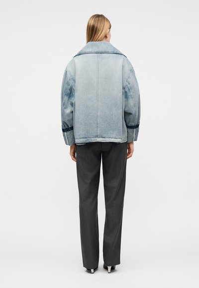 Frau mit glattem blondem Haar steht mit dem Rücken zum Betrachter, trägt eine hellblaue übergroße Jeansjacke, dunkelgraue gerade geschnittene Hosen und schwarze Schuhe mit Absatz.