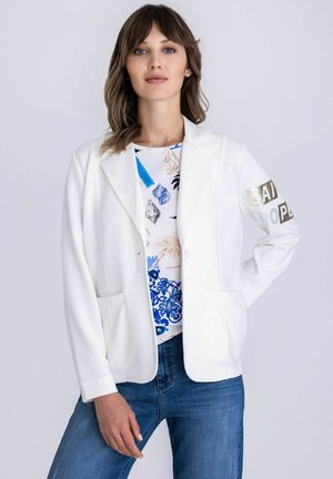 Vrouw met een wit colbert met tekst op de mouw, een wit blouse met patroon en een blauwe spijkerbroek, staand met een hand op haar heup.