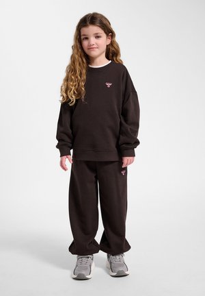 Jeune fille aux longs cheveux bouclés portant un sweat-shirt noir ample et un pantalon avec des logos d'abeilles roses, debout devant un fond uni.