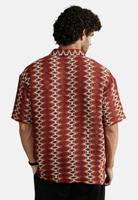 Camisa de manga corta en un patrón geométrico rojo y naranja con acentos en blanco, con cuello, hecha de una tela texturizada. Presenta un ajuste relajado.