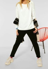 Top blanco de manga larga, pantalones negros y zapatillas beige. El top tiene un corte relajado, y los pantalones tienen una textura suave. Un chal estampado añade detalle.