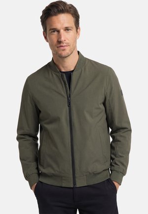 Homme portant une veste bomber vert olive avec une chemise noire en dessous, debout les mains dans les poches devant un fond uni.