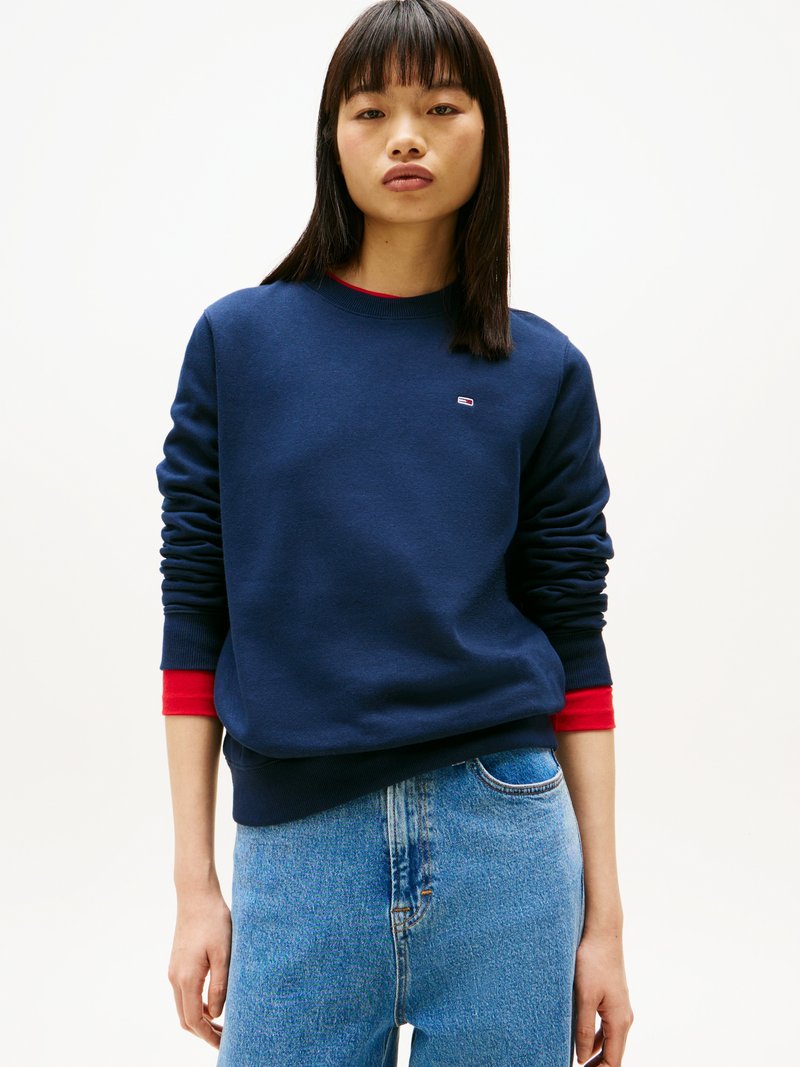 Sweatshirt bleu marine avec des accents rouges aux poignets, design à col rond, texture douce, porté sur un jean taille haute bleu clair. Logo présent.