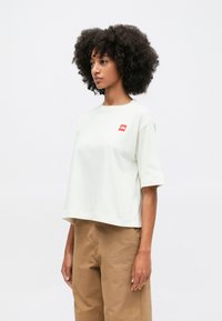 Heledabeige lühikeste varrukatega cropitud t-särk, ümmarguse kaelusega ja vasakul rinnal väike punane logo. Kanga pind näib pehme ja sile.