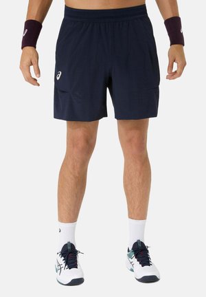 Man draagt marineblauwe sportshorts, witte sportschoenen met zwarte veters, witte enkelsokken en donkere polsbanden, staat met gespreide benen.